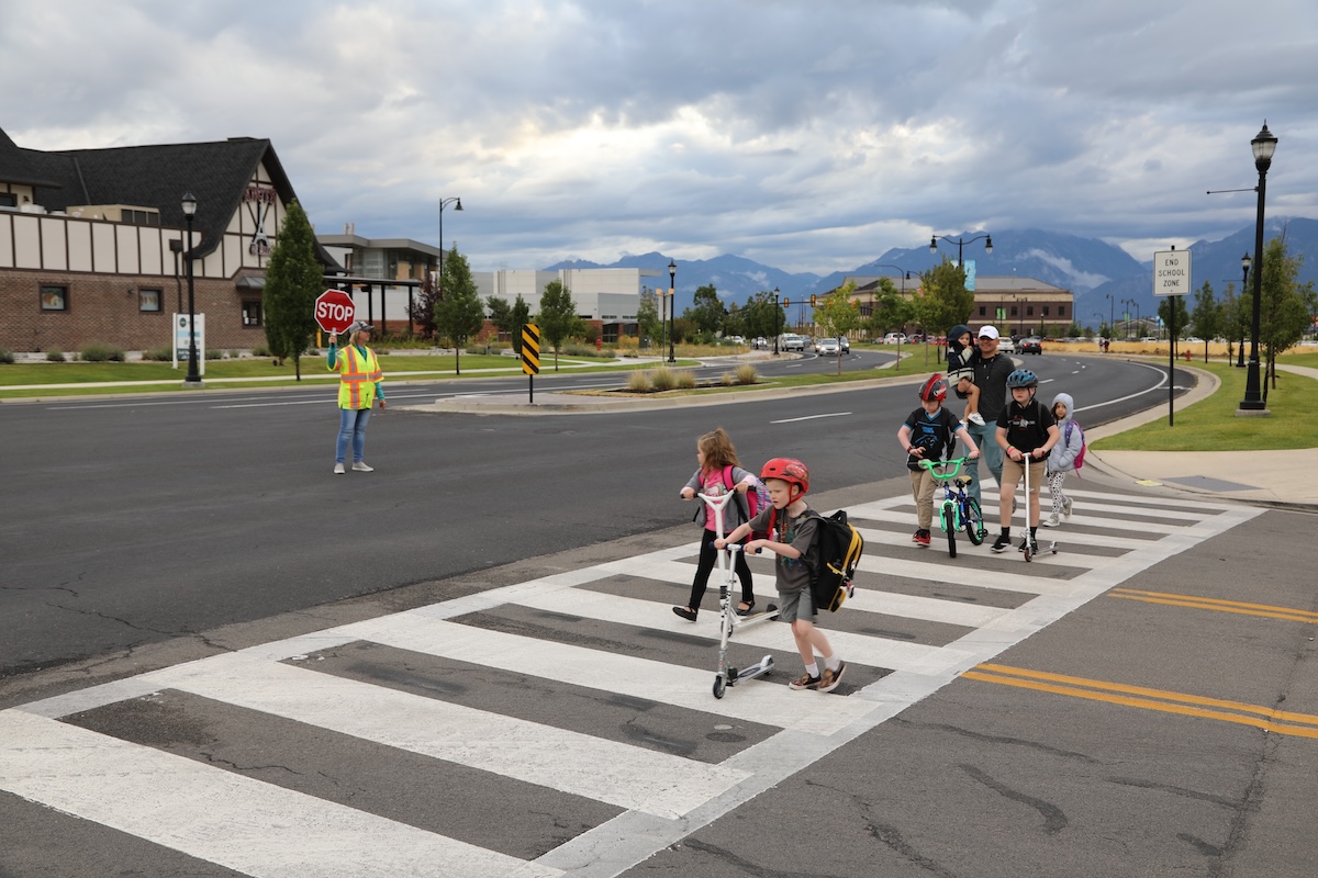 Children using scooters and bikes cross the street at the Herriman Rose Boulevard east-west crosswalk, guided by a crossing guard holding a stop sign. The crossing guard is wearing a reflective safety vest. The background shows a residential area with buildings, greenery, and mountains under a cloudy sky.